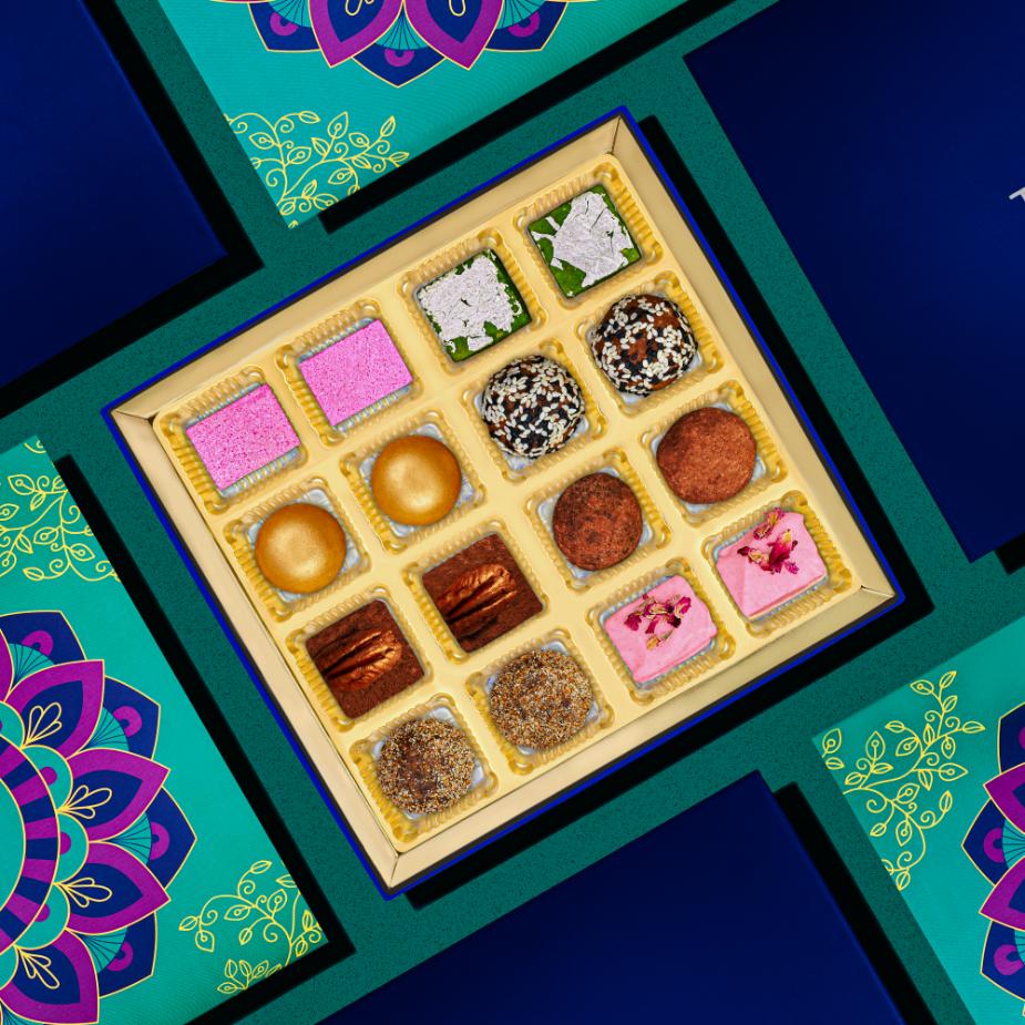 
                      
                        Colorful Diwali Mithai Collection box featuring 16 assorted Indian sweets in vibrant colors, displayed on a festive blue and green background with decorative floral patterns
                      
                    