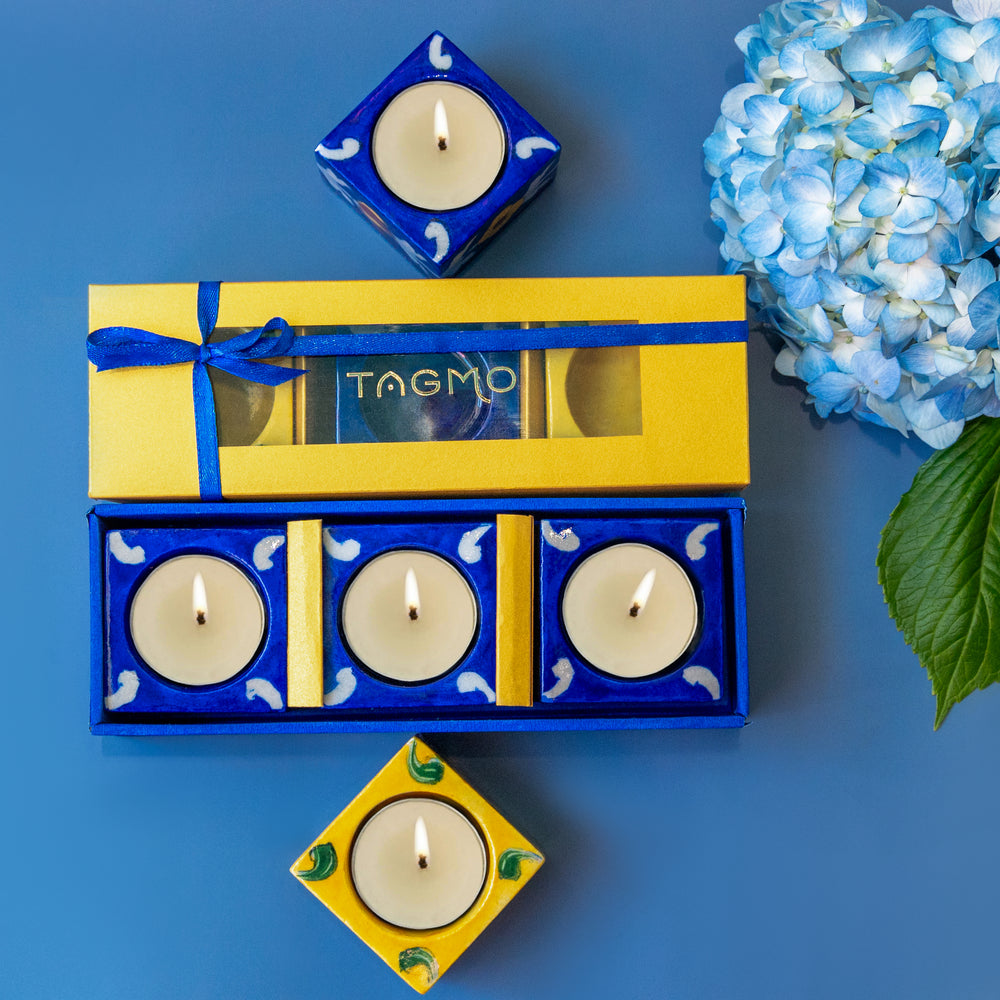 
                      
                        Hand-painted blue and yellow porcelain diyas in a gift box, displayed with lit candles and a blue hydrangea flower on a vibrant blue background. Artisanal craftsmanship from Jaipur showcased in a 3-pack set of decorative oil lamps.
                      
                    