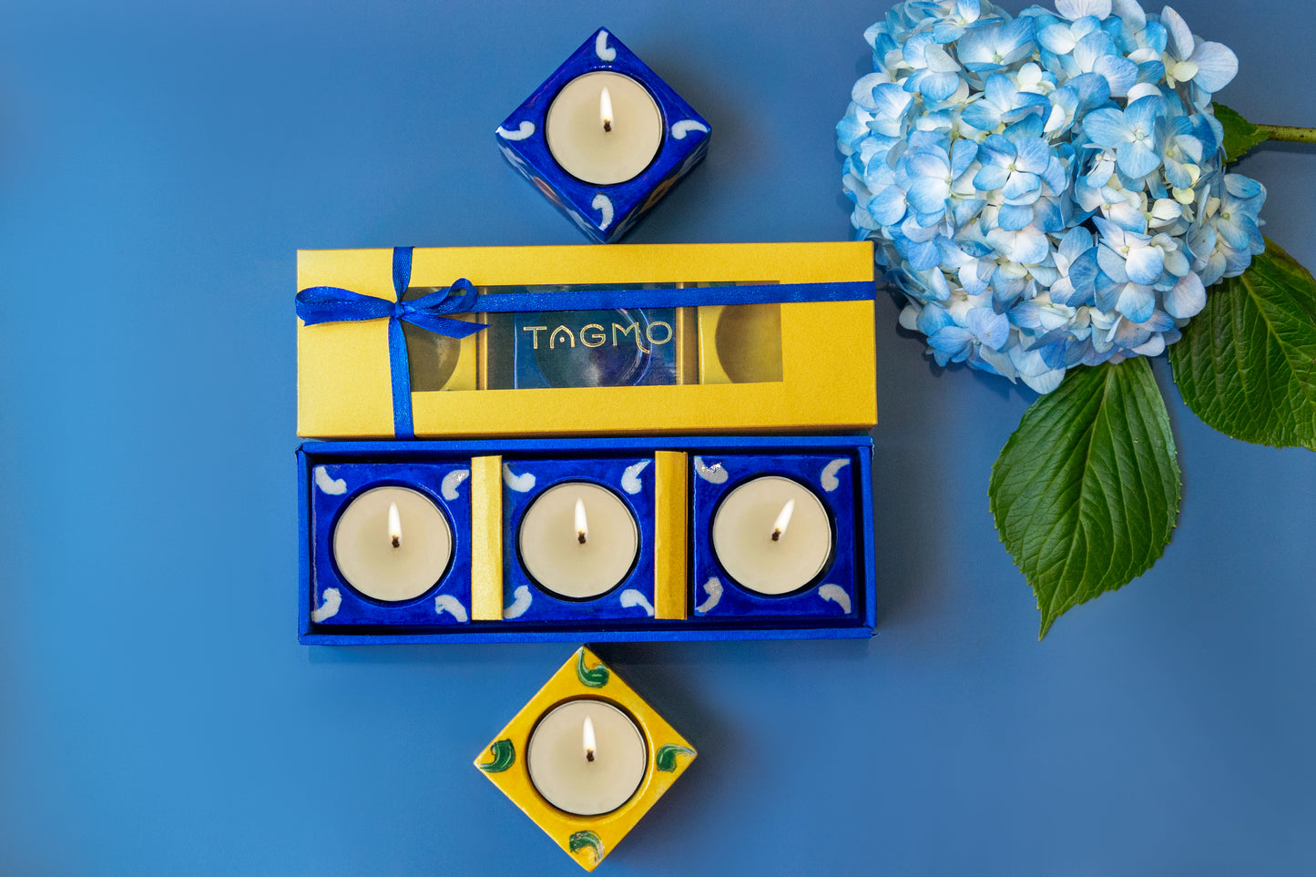 Hand-painted blue and yellow porcelain diyas in a gift box, displayed with lit candles and a blue hydrangea flower on a vibrant blue background. Artisanal craftsmanship from Jaipur showcased in a 3-pack set of decorative oil lamps.