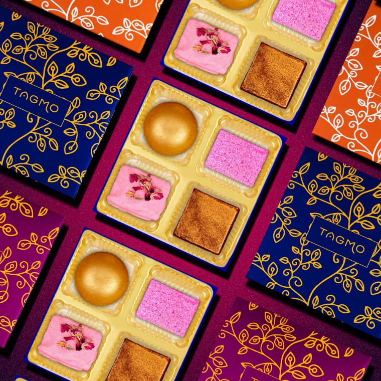 
                      
                        Colorful assortment of TAGMO favor boxes featuring vibrant Indian sweets and decorative packaging. Each box contains four compartments with different mithai varieties, including golden, pink, and brown treats, set against a rich purple background.
                      
                    