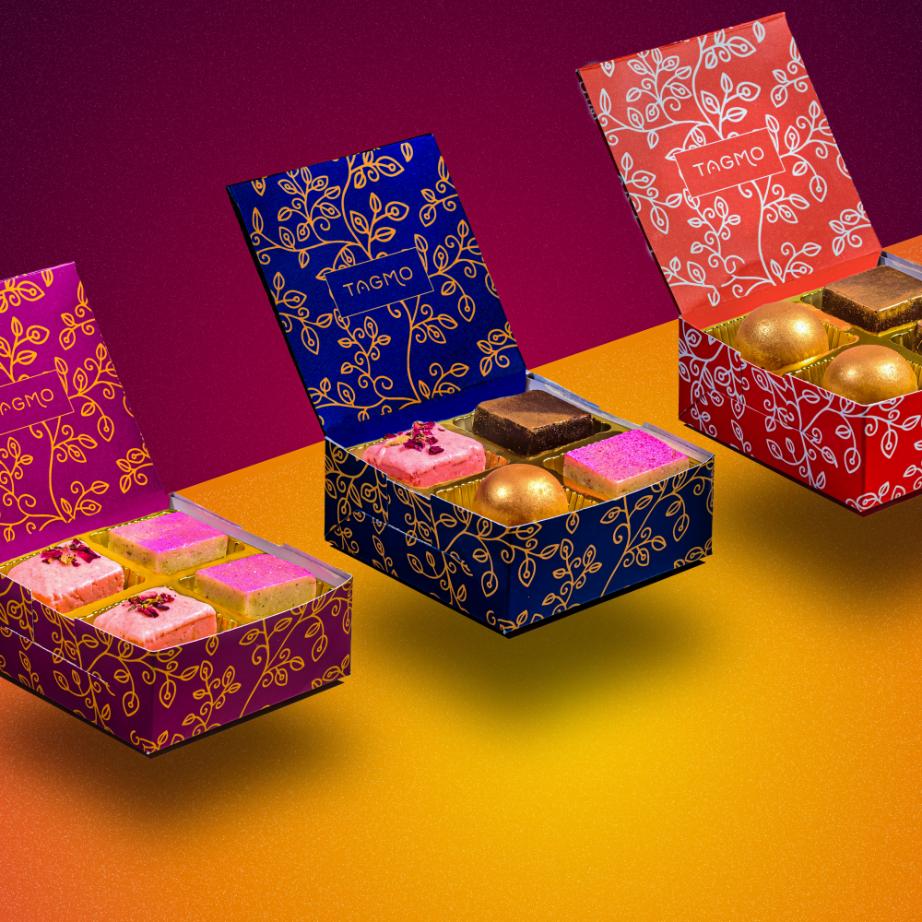 Colorful TAGMO favor boxes showcasing assorted Indian sweets, including burfi and ladoo, arranged on vibrant yellow and red background. Elegant floral patterns adorn the boxes, displaying a variety of flavors and designs.