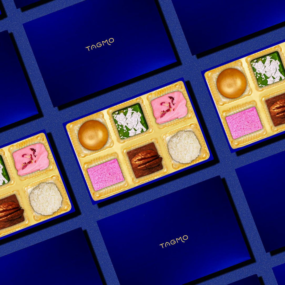 
                      
                        Elegant TAGMO favor boxes showcasing assorted colorful mithai treats in compartments, including pink, white, and brown sweets, arranged on a deep blue background for luxurious presentation
                      
                    