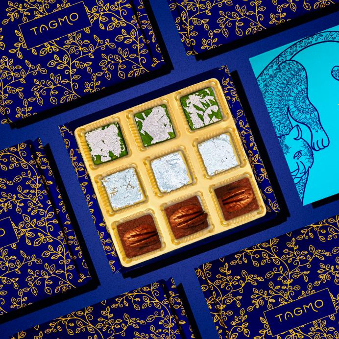 
                      
                        Assorted Indian burfi sweets in elegant gold and blue TAGMO gift boxes, featuring pistachio, kaju, and pecan varieties arranged in a visually appealing grid pattern.
                      
                    