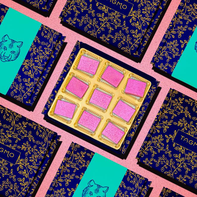 
                      
                        Colorful display of TAGMO's Kaju Katli box featuring pink-dusted diamond-shaped sweets, surrounded by patterned navy and teal packaging, arranged on a pink background
                      
                    