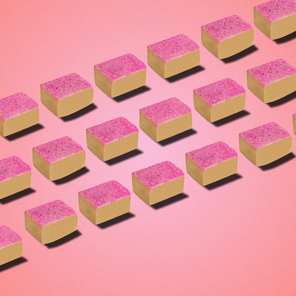 
                      
                        Square-shaped Kaju Katli sweets arranged in diagonal rows on a pink background. Each piece has a golden base and shimmering pink top, showcasing the unique TAGMO twist on this classic Indian mithai dessert.
                      
                    