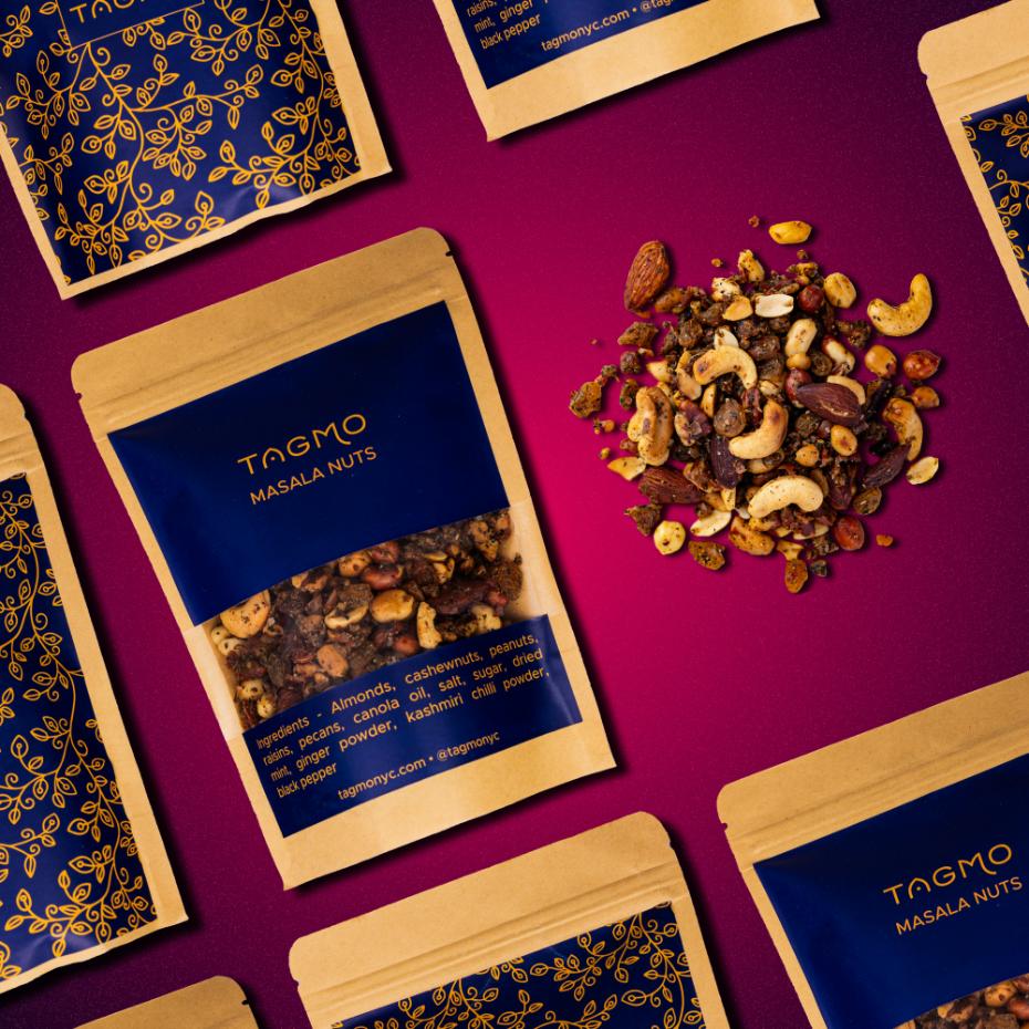 
                      
                        Tagmo's Masala Nuts product display featuring kraft paper packaging with blue labels, surrounded by ornate blue and gold gift boxes on a vibrant purple background. A heap of colorful mixed nuts and spices showcases the product's enticing contents.
                      
                    