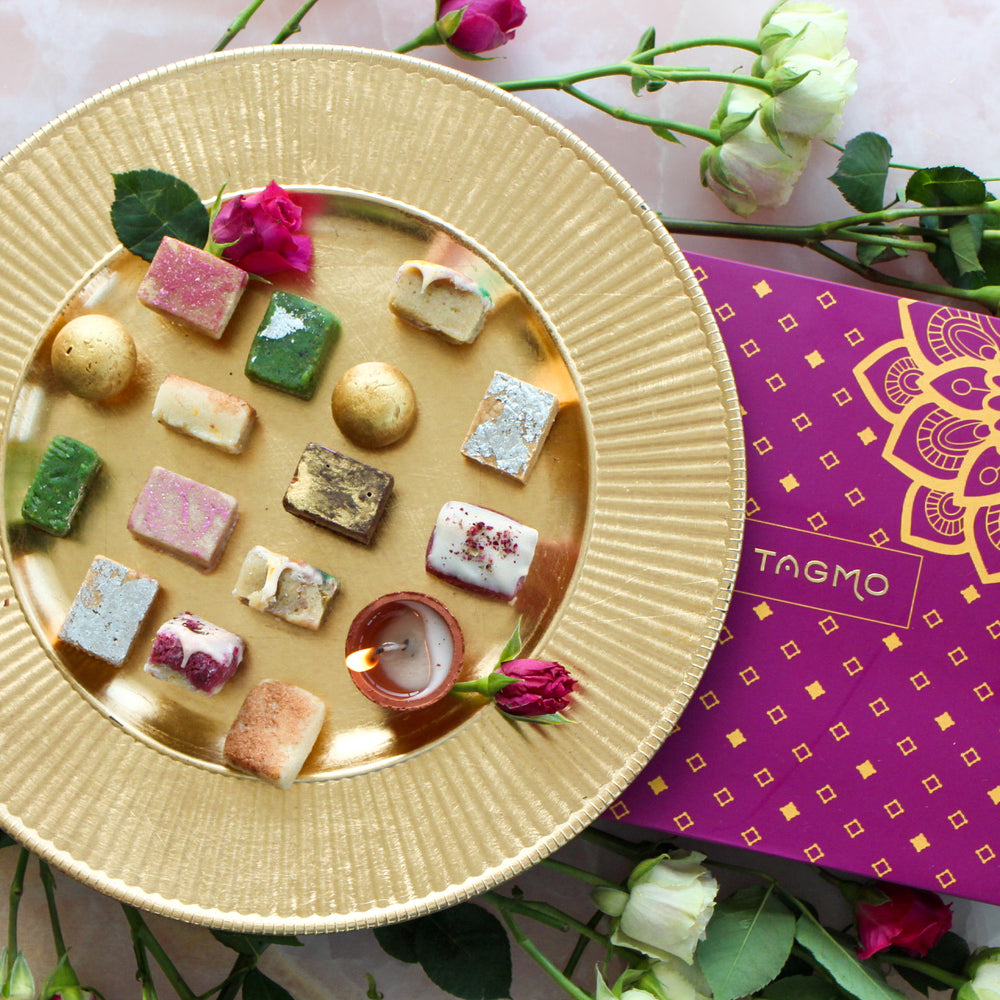 A colorful assortment of traditional Indian sweets, or mithai, arranged in an elegant gold dish, alongside a vibrant purple and gold box, creating a visually appealing and enticing display for the "Build Your Own Mithai Box" Shopify product.