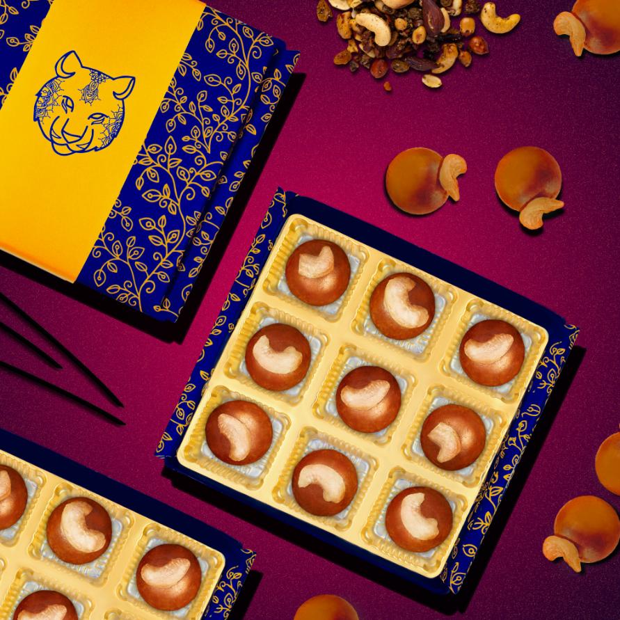 Colorful display of Nani Ke Ladoo boxes with golden ladoos arranged in elegant blue and yellow packaging, surrounded by assorted nuts and decorative elements on a deep purple background, showcasing traditional Indian sweets with a modern twist.