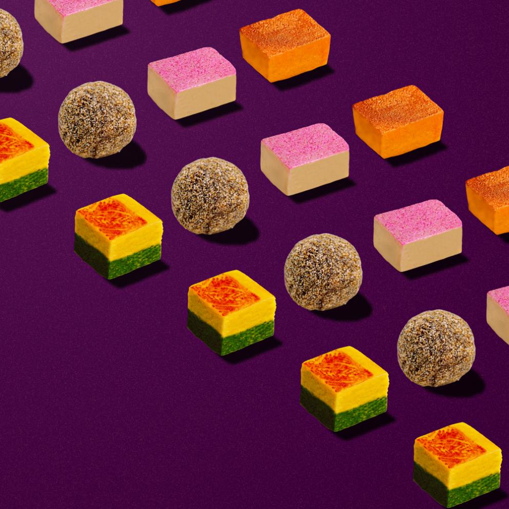 Colorful array of vegan Indian sweets on purple background, featuring square, rectangular, and round shapes in vibrant hues of pink, orange, green, and brown, showcasing the variety in TAGMO's Vegan Mithai Collection