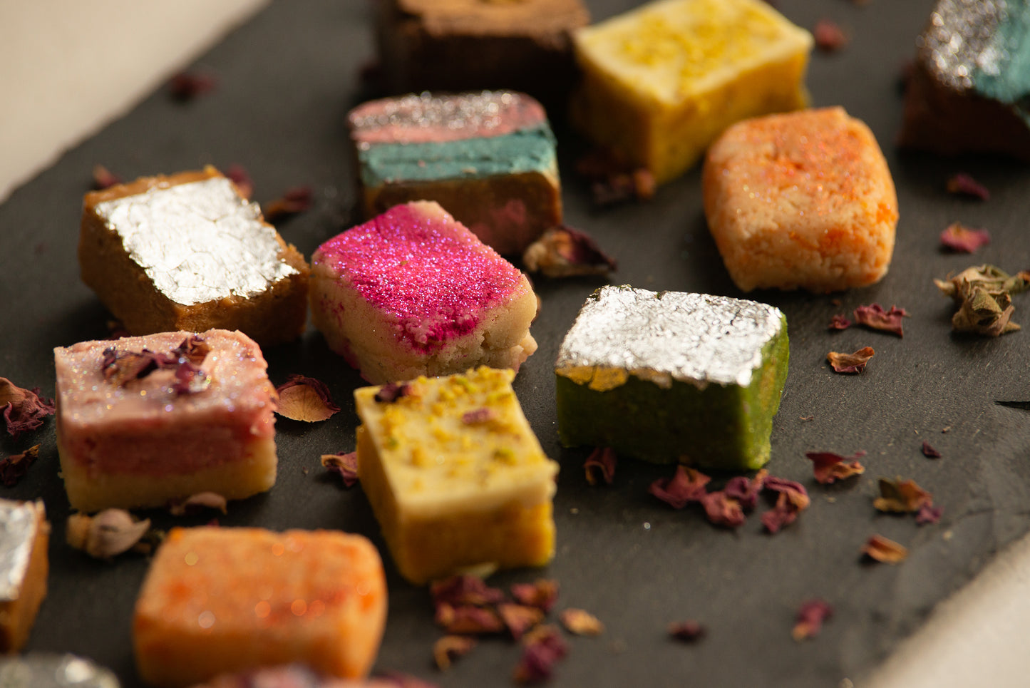 Colorful assortment of Indian sweets on a dark plate, including cubes with vibrant toppings like pink, white, and yellow, alongside scattered flower petals and spices, showcasing the variety of a custom mithai box